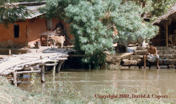 A man sits beside the water, in his village, reflecting.